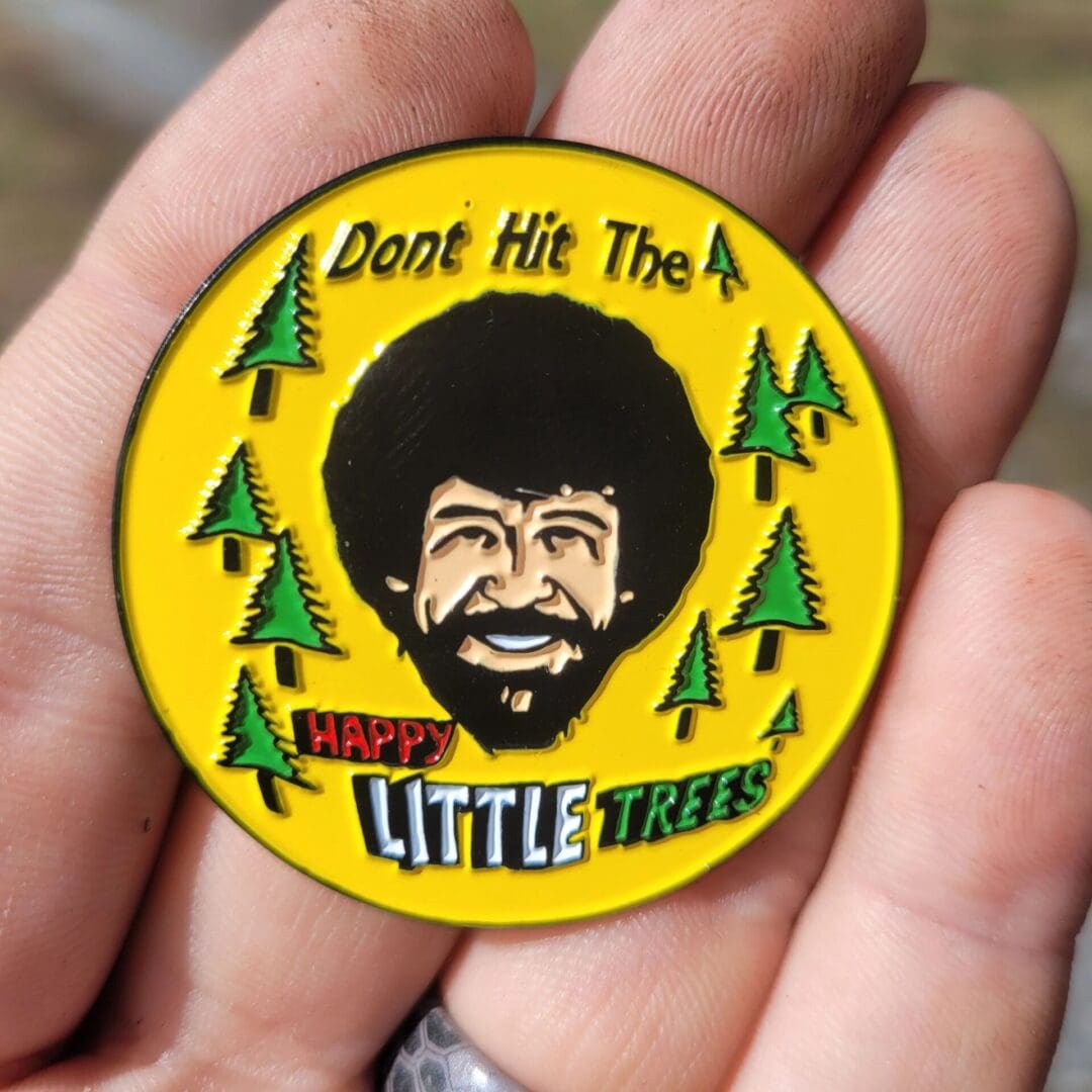 A person holding a pin with trees and a picture of bob ross.